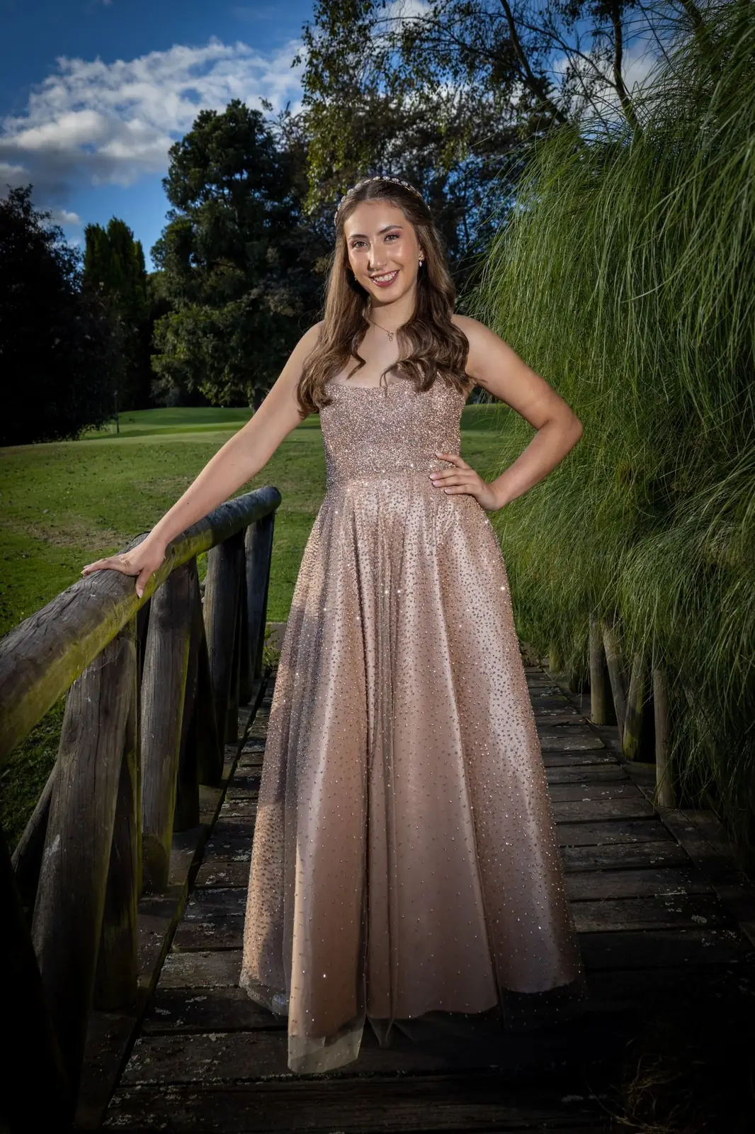 Quinceañera con vestido brillante en un puente de madera con cielo azul